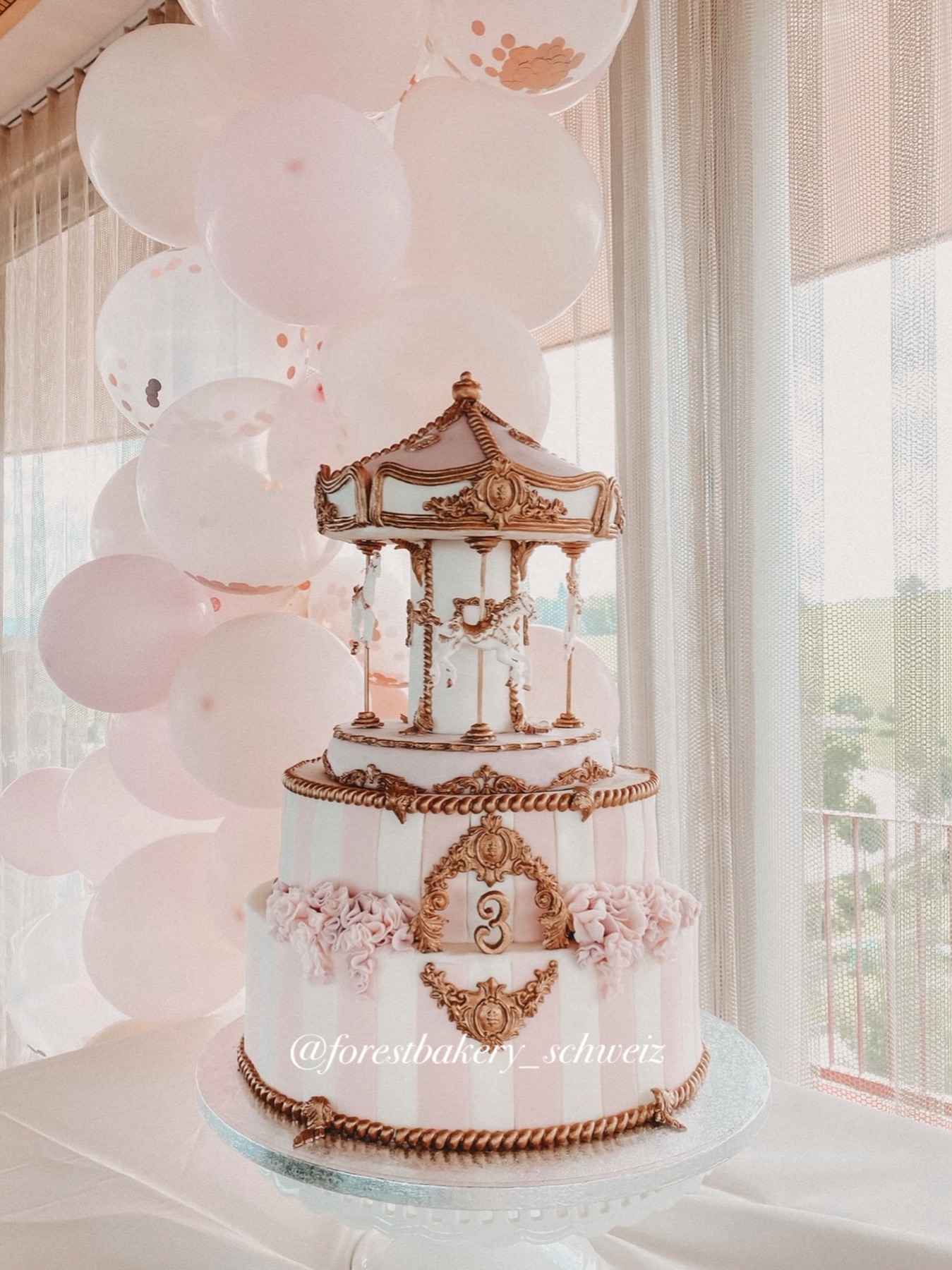 Rosa-weisse Karusseltorte mit goldenen Verzierungen vor rosa Ballonen